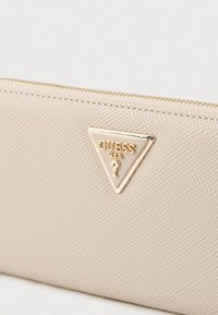 Beige synthetische portemonnee met een textuur oppervlak, een gouden driehoekig logo "GUESS USA" en een ritssluiting langs de bovenkant.