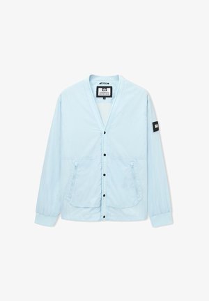 Veste boutonnée bleu clair à manches longues, deux poches zippées à l'avant, poignets élastiques et un patch noir sur la manche gauche.