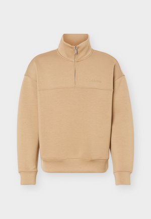 Béžová mikina s půlzipem, vysokým límcem, dlouhými rukávy, žebrovanými manžetami a lemem a nenápadným logem značky na hrudi.