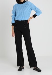 Ljust blå stickad tröja med ett strukturerat mönster, svarta detaljer, högmidjade svarta byxor med knappdetalj fram, och svarta loafers.