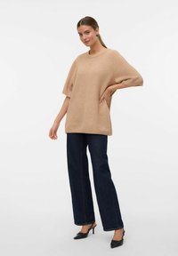 Beige übergroßer Strickpullover mit kurzen Ärmeln, Rundhalsausschnitt und seitlichen Schlitzen, kombiniert mit geraden dunkelblauen Jeans und schwarzen Absatzschuhen.