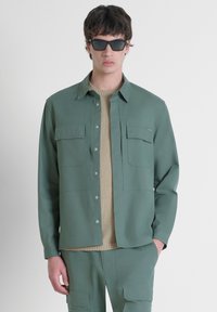 Camicia verde con bottoni e due tasche sul petto, indossata sopra un maglione beige lavorato a maglia. Il modello indossa anche occhiali da sole neri a forma quadrata.