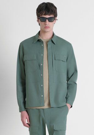 Groene button-up shirt met twee borstzakken, gedragen over een beige gebreide trui. Model draagt ook zwarte vierkante zonnebril.