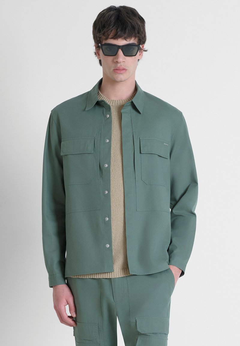 Camicia verde con bottoni e due tasche sul petto, indossata sopra un maglione beige lavorato a maglia. Il modello indossa anche occhiali da sole neri a forma quadrata.