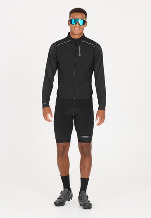 Schwarze Radjacke mit Reißverschluss, ausgestattet mit reflektierenden Akzenten, kombiniert mit schwarzen gepolsterten Shorts und Radschuhen. Große blaue Sonnenbrille rundet den Look ab.