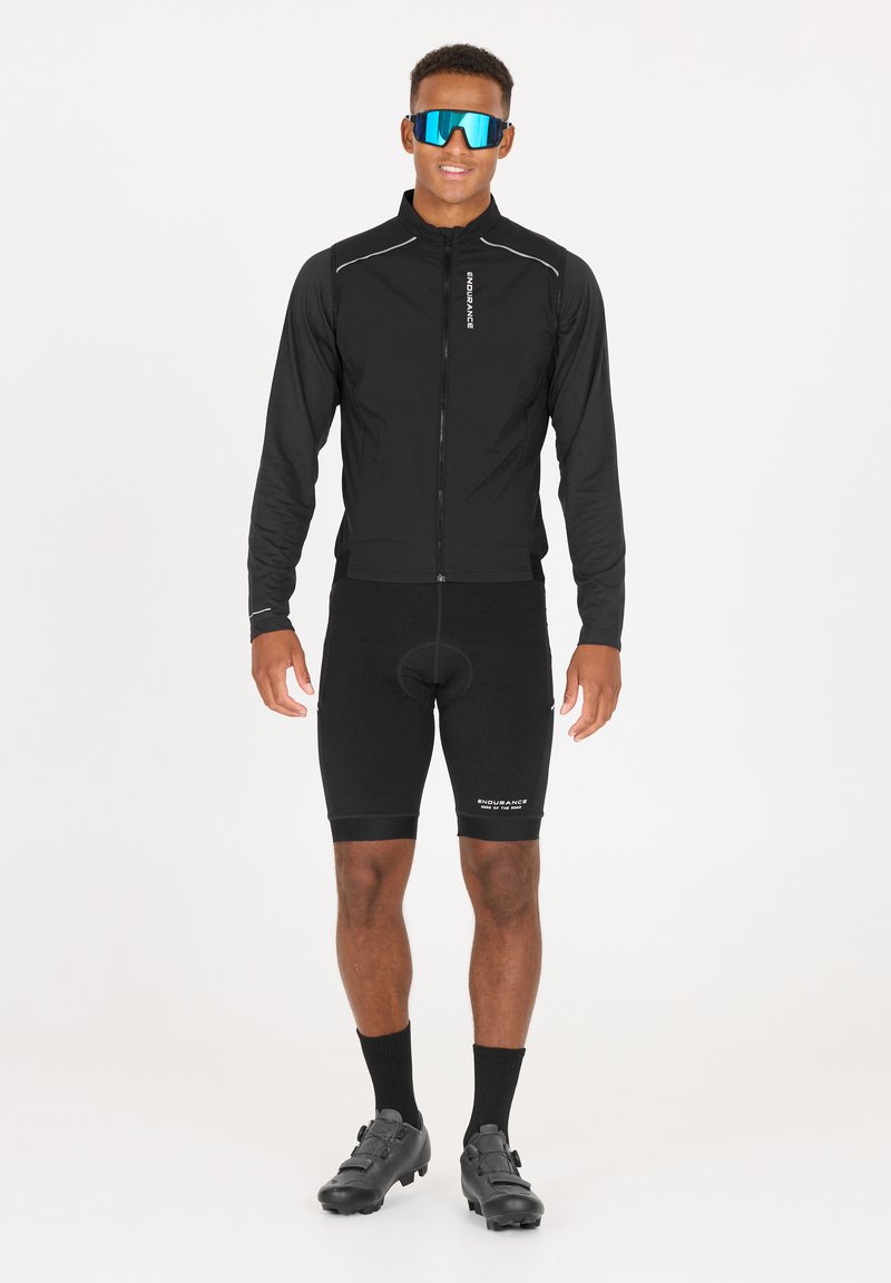 Schwarze Radjacke mit Reißverschluss, ausgestattet mit reflektierenden Akzenten, kombiniert mit schwarzen gepolsterten Shorts und Radschuhen. Große blaue Sonnenbrille rundet den Look ab.