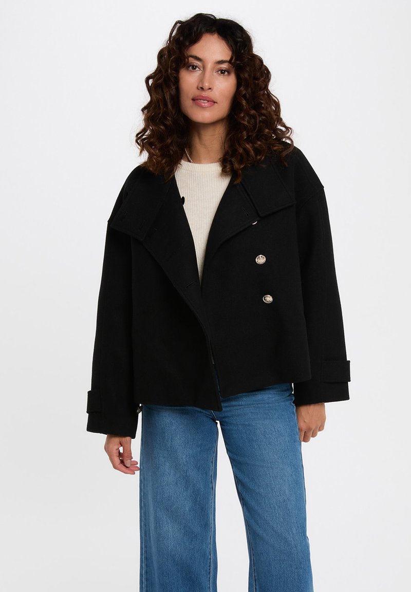 Schwarze doppelt geknöpfte Jacke mit großen Revers, silbernen Knöpfen und weiten Ärmeln, kombiniert mit blauen Jeans und einem cremfarbenen Top.