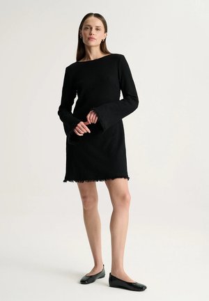 Kobieta stojąca, ubrana w czarną sukienkę z długimi rękawami z rozszerzanymi mankietami oraz czarne baleriny na jednolitym, jasnym tle.