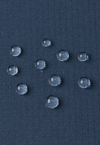 Il tessuto impermeabile presenta chiare gocce d'acqua che si adagiano su una superficie strutturata di colore blu scuro, evidenziando la resistenza all'acqua e la durabilità.