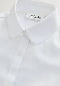 Camicia bianca in un tessuto liscio, con colletto classico a punta, chiusura con bottoni e un'etichetta con "Clarks" cucita all'interno del colletto.
