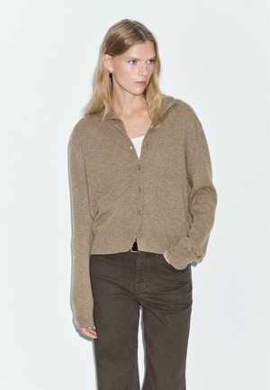 Cardigan beige clair avec col, featuring un fermeture à boutons, des poignets côtelés et une coupe courte, associé à un pantalon marron foncé.