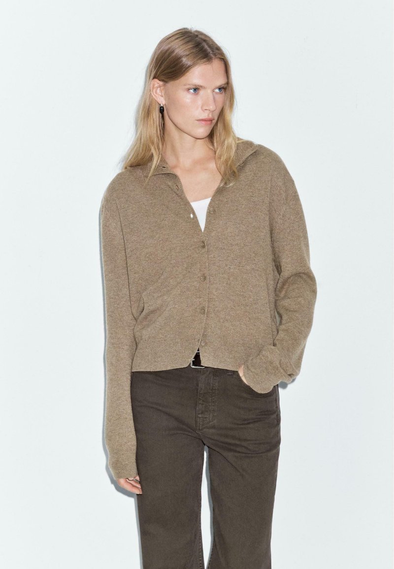Light brown cardigan with a collar, featuring button closure, ribbed cuffs, and a cropped design, paired with dark brown trousers.