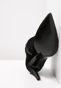Zapato de tacón alto negro hecho de tela texturizada con punta afilada y tacón negro brillante. Detalle de cierre con cremallera lateral visible.