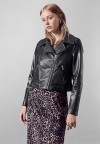 Giubbotto da motociclista in pelle nera con ampio colletto, chiusura con zip frontale e tasche laterali, abbinato a un vestito con stampa leopardo. Texture liscia.