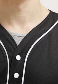 Maillot de baseball noir en tissu en maille, avec des piping blancs le long du col et de la patte de boutonnage. Trois boutons blancs sont visibles.