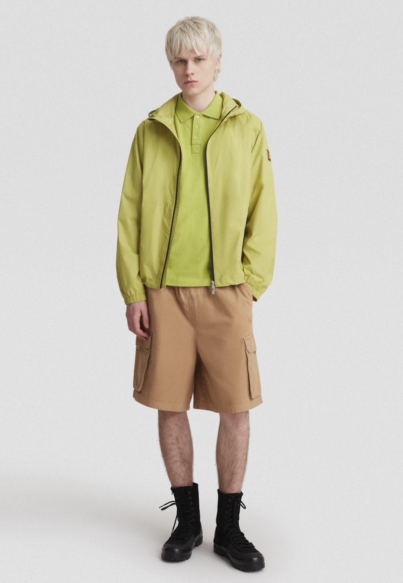 Hellgrüne Zip-Jacke über einem passenden Polo, kombiniert mit hellbraunen Cargo-Shorts und schwarzen Stiefeln. Glatte Materialien und entspannte Passform.