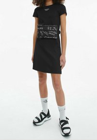 Černé šaty s krátkými rukávy a přiléhavým střihem, které mají odnímatelnou kabelku v pase černé barvy s bílými logy. Kombinováno s bílými ponožkami a sandály.