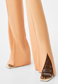 Peach wide-leg pants with side slits and smooth texture, paired with transparent heeled sandals featuring a squared toe design.