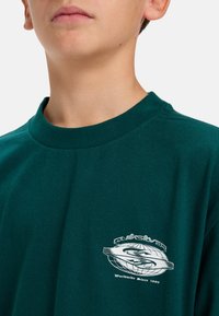 Zeleni pamučni majica s okruglim izrezom i kratkim rukavima, s bijelim logotipom na području prsiju i glatkom teksturom.