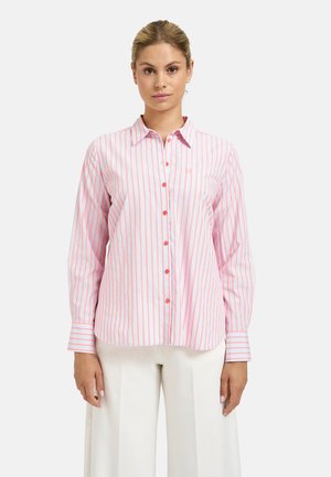 Woman wearing a long-sleeve button-up shirt with pink vertical stripes and white wide-leg pants, standing against a plain background.