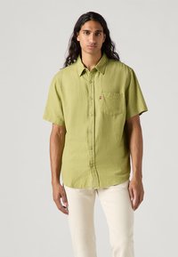Camicia a maniche corte con bottoni in tessuto testurizzato verde chiaro, dotata di taschino sul petto e branding discreto. Abbinata a pantaloni chiari.