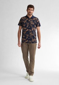 Marinblå polo tröja med ett blommönster i beigefÄrger, kombinerad med ljusbruna byxor och vita sneakers. Korta ärmar, klassisk krage.