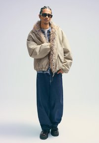 Bézs bombázó dzseki műszőrme gallérral, kockás ing felett viselve, laza sötét farmernadrággal és vastag fekete cipőkkel párosítva.