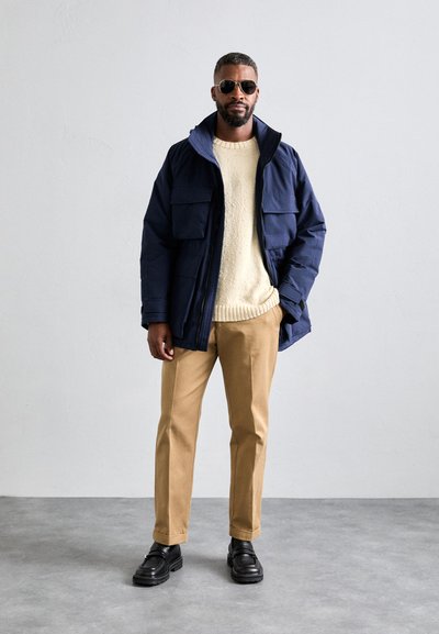 Giacca imbottita blu navy con diverse tasche frontali, maglione testurizzato crema, pantaloni beige e scarpe nere chunky. Uomo con occhiali da sole.