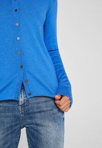 Cardigan blu realizzato in tessuto morbido con maniche lunghe, presenta una chiusura frontale con bottoni e polsini a costine. Abbinato a jeans in denim consumati.