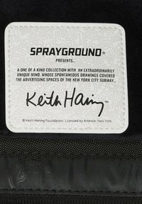 Etiqueta rectangular blanca con un patrón en relieve y texto negro. El texto incluye "SPRAYGROUND," "Keith Haring," y una declaración de licencia en la parte inferior.