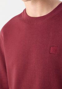 Gros plan d'une personne portant un sweat-shirt à col rond bordeaux avec un petit logo carré embossé sur la poitrine gauche.