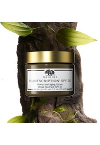 Grønn glasskrukke med gullokk, merket "Plantscription SPF 25," som inneholder anti-aldringskrem, omgitt av blader på teksturert tre.