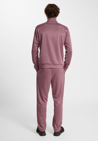 Pullover træningsdragt sæt i afdæmpet lyserød, lavet af glat stof. Har en høj krave og elastiske manchetter ved ærmerne og buksekanterne.