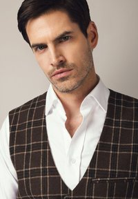 Brązowa kamizelka w kratkę noszona na białej koszuli, z kołnierzykiem typu notch i małą kieszonką po lewej stronie, o gładkim i dopasowanym kroju.