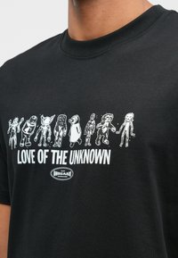 Czarna koszulka z białą grafiką ośmiu postaci obcych nad napisem "MIŁOŚĆ DO NIEZNANEGO" oraz małym logo "NØRGAARD" poniżej.