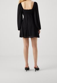 Svart miniklänning med fyrkantig halsringning, puffärmar och smockad baksida. Det genomskinliga tyglagret ger en flödande siluett. Bärs med svarta klackar.