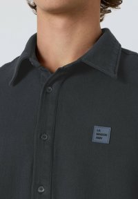 Zblizu pogled na osebo, ki nosi temnosiv srajčni gumbasti vzorec s majhnim kvadratnim narekovajem "LA MAISON AMV" na prsih.