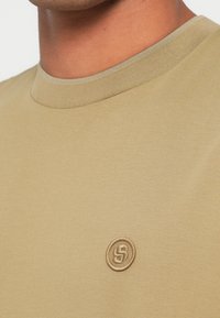 Oliwkowa koszulka z okrągłym dekoltem, wykonana z gładkiej tkaniny, z małym, haftowanym logo na lewej piersi.