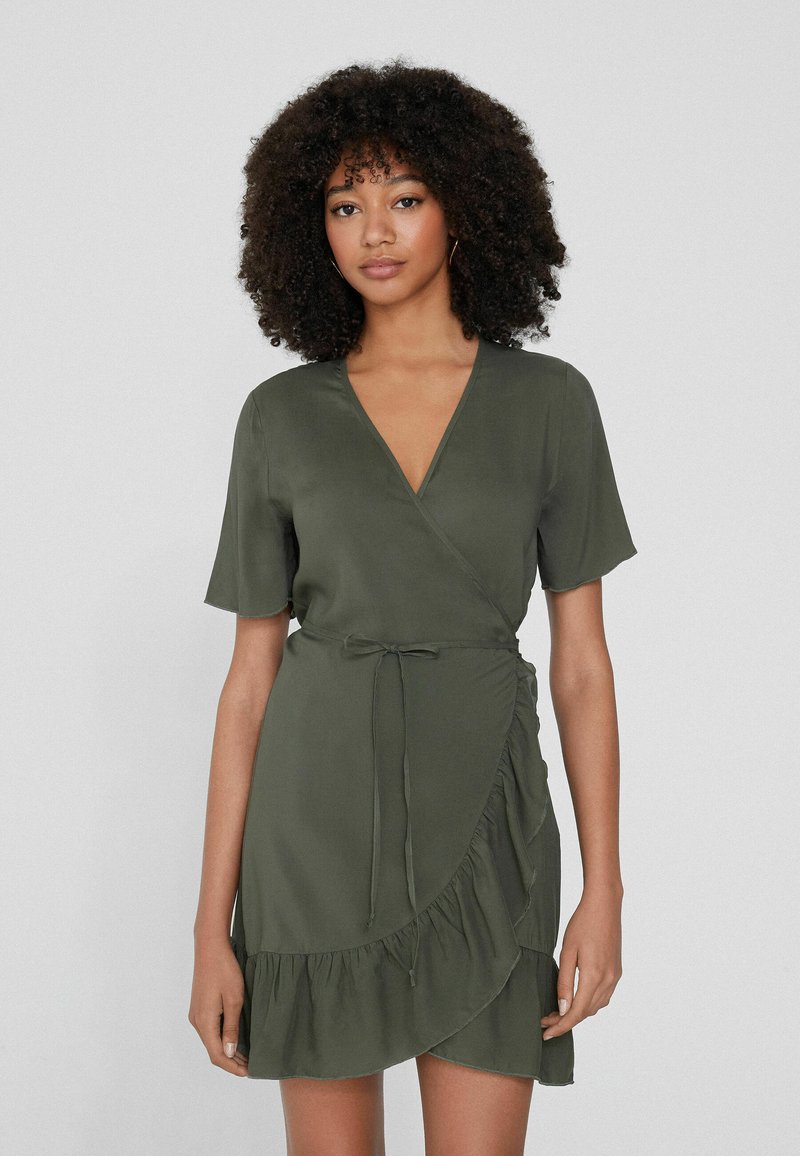 Vestido de envoltura verde oliva, de manga corta, con escote en V, cintura ajustada con lazo y dobladillo con volantes. Tela suave y ligera con un diseño fluido.