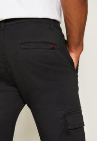 Zwarte cargo broek heeft een zachte textuur, een zijzak en een klein rood en marineblauw logo detail nabij de achterzak.