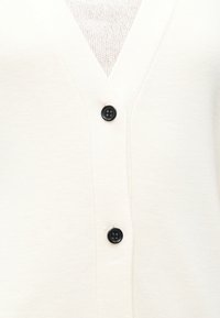 Λευκό πλεκτό cardigan με V λαιμόκοψη, δύο μαύρα κουμπιά και ένα υφασμάτινο εσώρουχο με υφή που είναι μερικώς ορατό από κάτω.