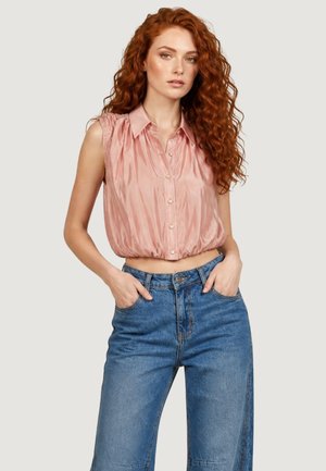 Femme aux longs cheveux roux bouclés portant un chemisier en satin rose sans manches et un jean bleu taille haute, les mains dans les poches, fond neutre.