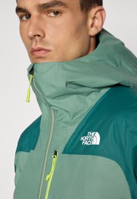 Veste zippée verte avec capuche, yoke vert foncé contrastant et logo réfléchissant. Le zip d'accent est équipé d'une tirette vert citron.