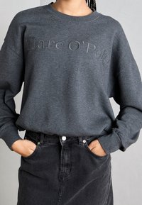 Felpa grigia con logo "Marc O'Polo" in rilievo. Presenta maniche lunghe e una vestibilità morbida, abbinata a una gonna di jeans nera.