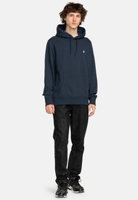 Maritiem blauwe hoodie met een kangoeroezak, capuchon met trekkoorden en een klein logo. Gecombineerd met zwarte jeans en gymschoenen.