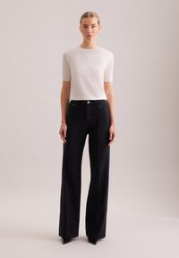 Maglia bianca a maniche corte abbinata a jeans neri a vita alta e gamba larga. I jeans presentano tasche anteriori e un dettaglio di bottoni a contrasto.