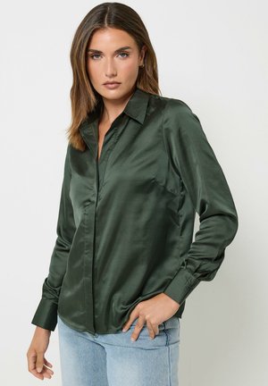 Vrouw met schouderlang bruin haar die een donkergroene satijnen blouse met lange mouwen en lichtblauwe spijkerbroek draagt tegen een effen achtergrond.