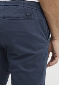 Mörkblå bomullsbukser med elastisk midja och sidofickor. En liten etikett syns på baksidan som anger märket.