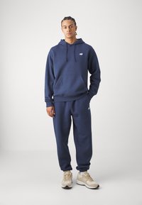 Marineblå hettegenser og matchende joggebukser laget av myk stoff, har en frontlomme og logo på brystet. Kombinert med beige treningssko.