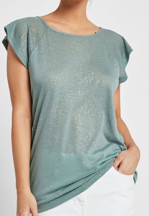 Mujer con una camiseta de manga corta verde claro con un sutil brillo plateado y pantalones blancos, con una mano en el bolsillo.